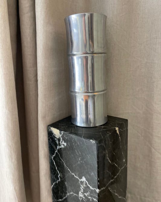Metallic duct on a black marble base against a beige curtain.