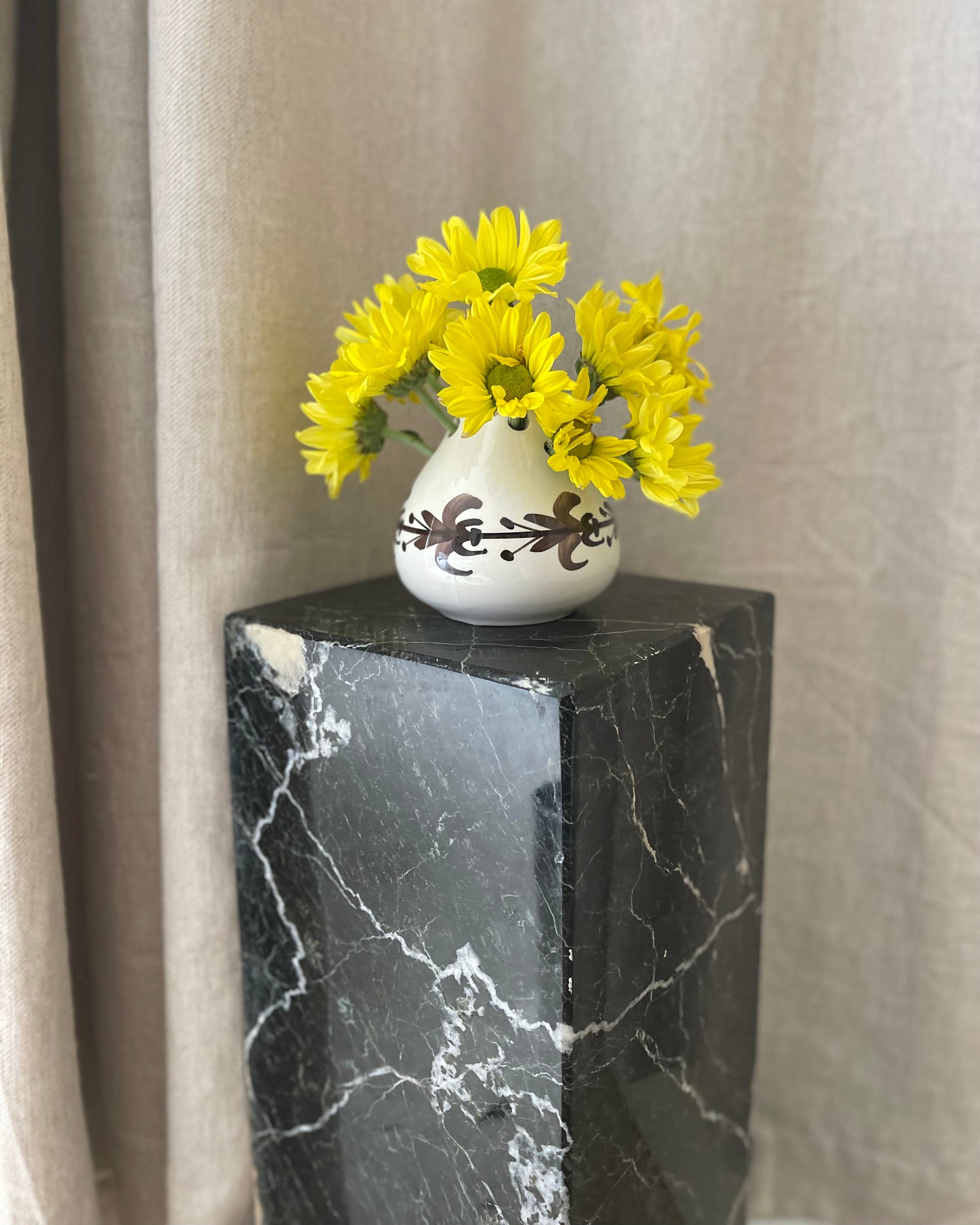 White vase with yellow flowers on a black marble pedestal against a beige curtain.