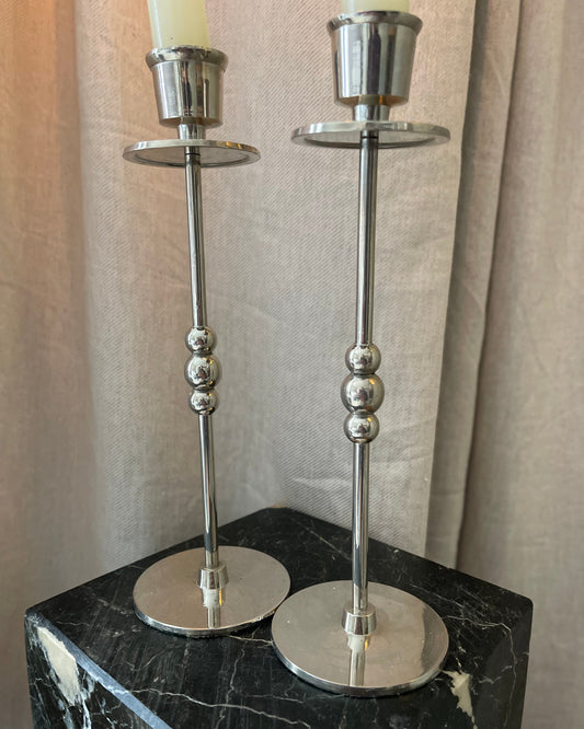 Two silver candle holders on a black marble surface with beige curtains in the background.