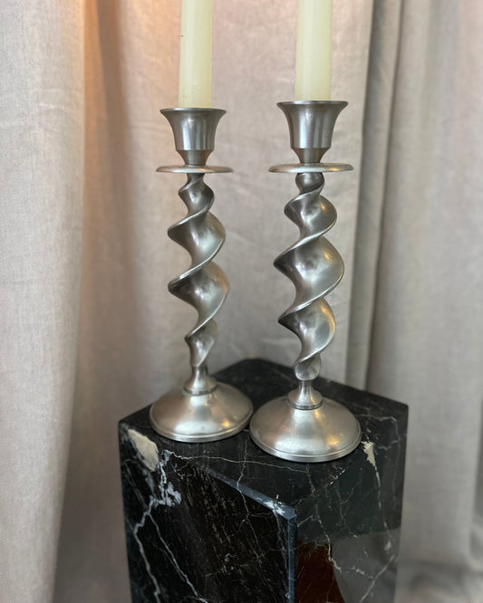 Two silver spiral candle holders with candles on a black marble stand against a neutral curtain background.